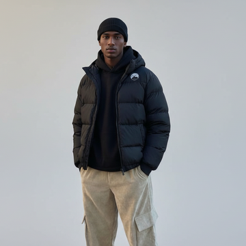 Person wearing a black puffer jacket, black beanie, and beige pants standing on a paved area with buildings in the background.